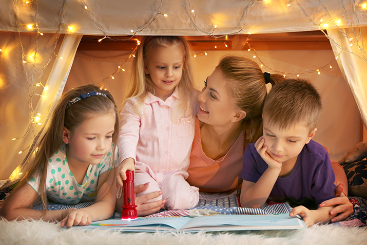 Image Description: A mom is laying on her stomach inside a tent. There are two kids to one side, and another on her other side. They are reading a book together. 