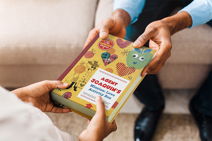 Image Description: A man's hands are holding an activity box. He is handing it to a child, whose hands are also holding the other side of the box.