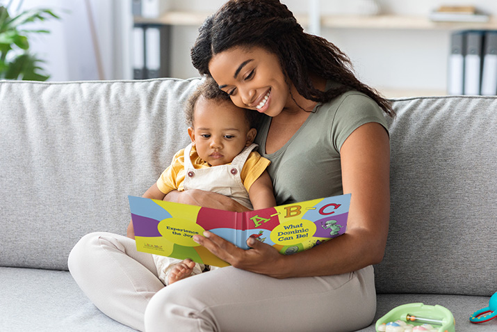 Image Description: A mom is sitting on a couch, holding open a storybook. Her other hand is wrapped around her baby. They are both looking at the book. 